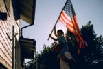 A father carrying his daughter on his shoulders as she raises an American flag to place on the outside of their house.