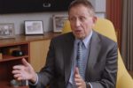 A person in a suit is seated in an office, speaking and gesturing with their hands. Framed photos are on the shelf in the background.